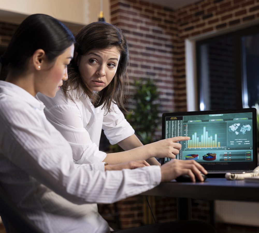 Diverse women collaborate and discuss strategy ideas in a late night office meeting, using financial graphs on laptop. The scene portrays a professional and focused atmosphere with dim lighting.